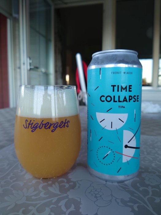 Ölburk med texten "Time Collapse TIPA" bredvid ett glas öl på ett bord utomhus.