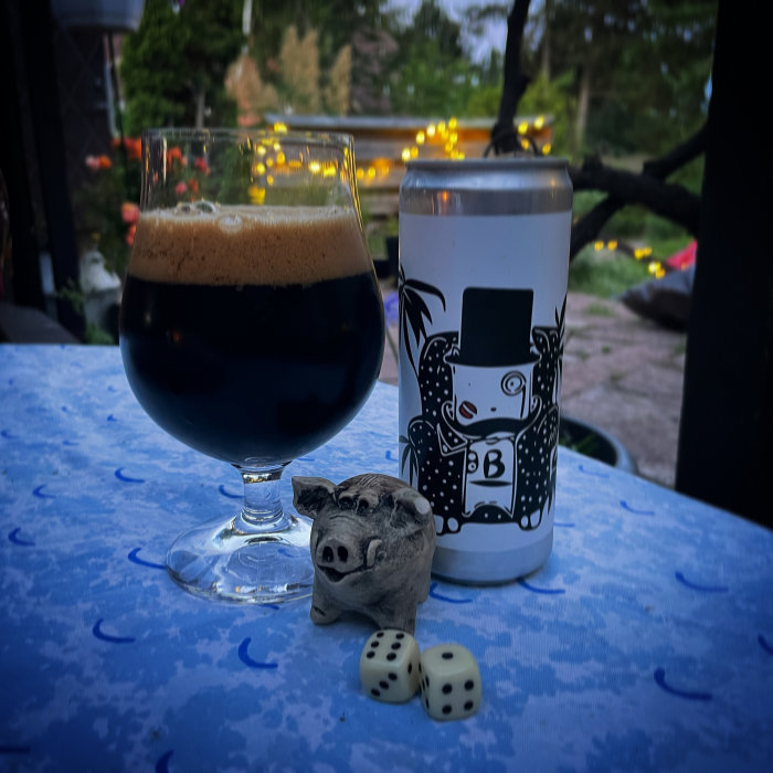 Ölglas med mörkt skummande öl, burk med konstnärlig design, träsvinsfigur och två tärningar på ett bord utomhus.