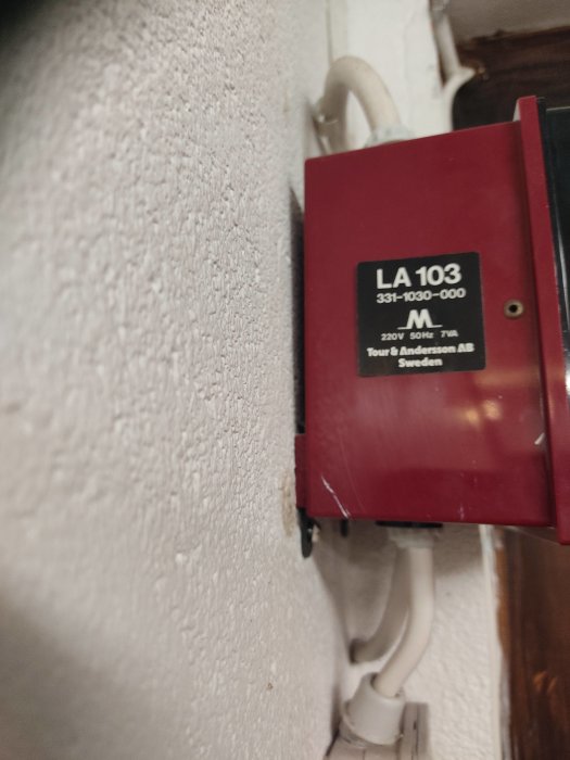 Röd äldre brytarbox märkt 'LA 103' monterad på vit vägg med kabel ansluten, misstänksam ljussensor.
