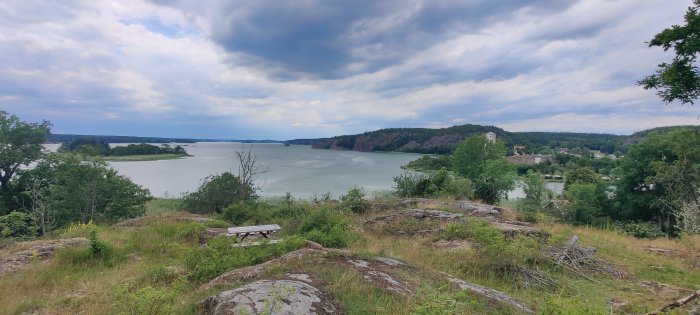 Enskild bänk med utsikt över ett lugnt vattendrag, omringad av skogklädda kullar och molntäckt himmel.