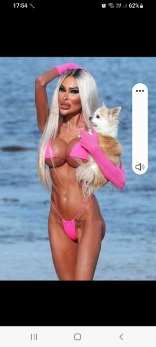 Kvinna i rosa bikini poserar på stranden med en liten hund i sina armar.