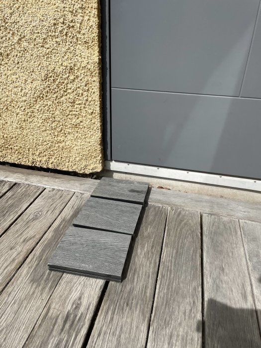 Ett prov av grått komposit-trä placerat på ett befintligt solblekt trätrall för jämförelse vid en husentré.