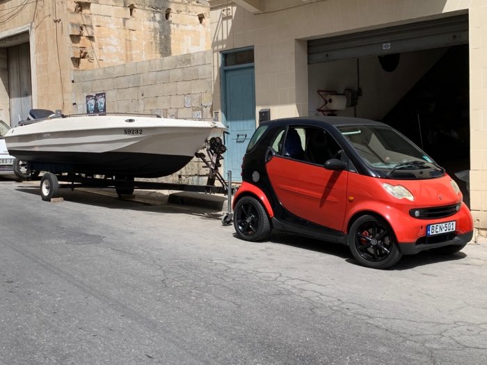 Liten röd bil knappt större än dess båttrailer parkerad på gatan.