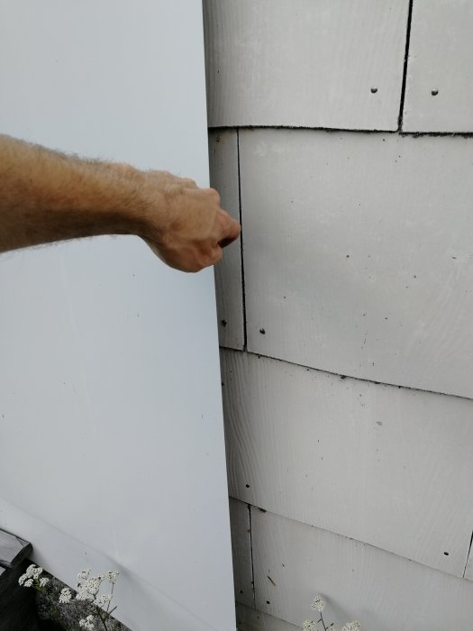 Hand som pekar på lösa skruvar i vit plåtmontering mot en eternitfasad med sprickor runt plåten.