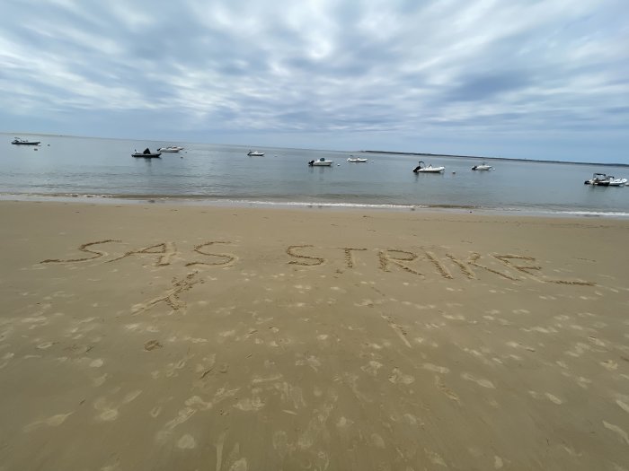 Strand med text skriven i sanden och båtar i det lugna havet i bakgrunden.