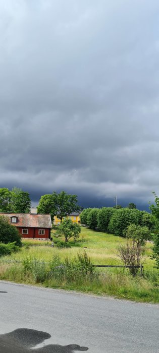 Ett landskap med ett rött hus, gul byggnad i bakgrunden och mörka moln på himlen.