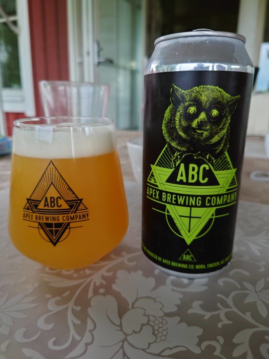 Ölglas med skummad ljusgul öl och en ölburk från Apex Brewing Company på mönstrad duk.