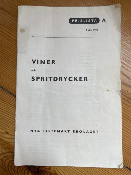 Gammal priskatalog från 1 oktober 1955 för viner och spritdrycker från Nya Systemaktiebolaget på ett träbord.