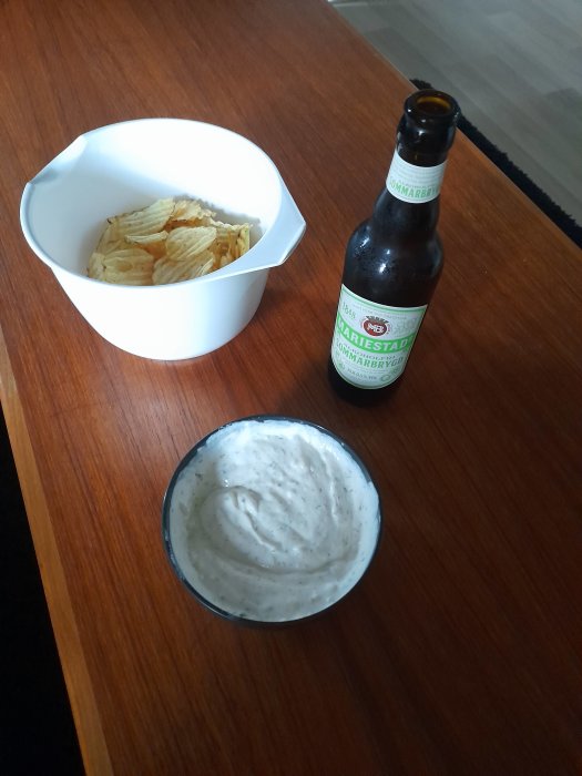 En skål med chips, en skål med dipp och en flaska öl på ett bord.