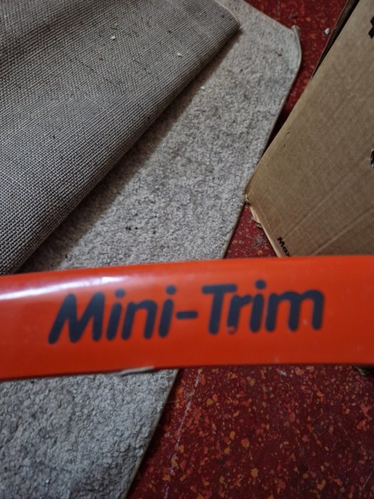 Röd föremål med texten "Mini-Trim" på en fläckig röd matta och en beige mattbit.