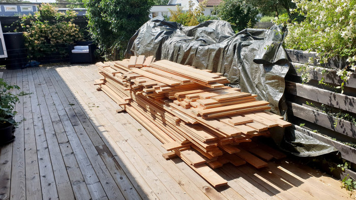 Staplar med avlånga träplank ordnade på ett trägolv utomhus, med presenningar och växtlighet i bakgrunden.