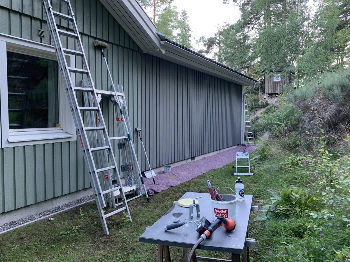 Stegar lutade mot husvägg i målningsprojekt med öppna färgburkar och verktyg på arbetsbord.