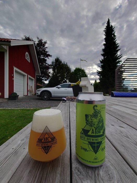 Apex Brewing Company ölburk och fyllt glas på träbord med hus och trädgård i bakgrunden.