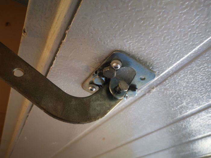 Närbild på en skadad infästning för en elektrisk portöppnare i ett vit plåtgargeport, med synligt slitage och skruvar.