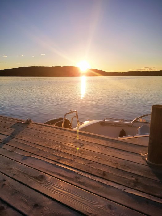 Solnedgång över en stilla sjö sedd från en träbrygga med en vit båt förtöjd vid kanten.