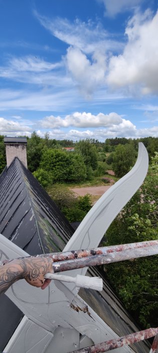 Person målar en vindskiva vit på ett hus med sluttande tak och utsikt över trädlanskap.