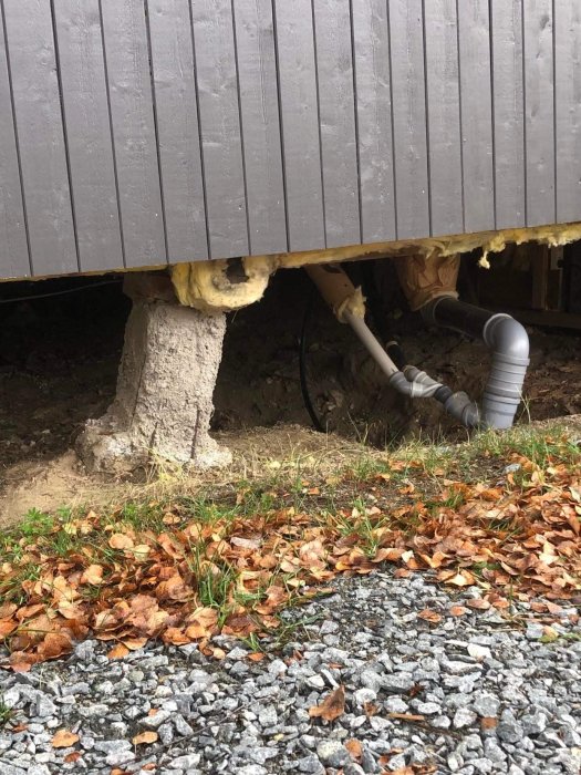 Bild på en lutande betongplint under hus med synlig isolering och rörledningar, med grus och löv i förgrunden.