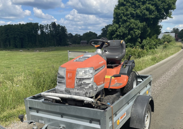 Husqvarna trädgårdstraktor lastad på en släpvagn parkerad vid en väg med äng och träd i bakgrunden.