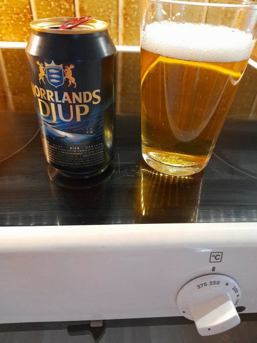 Burk av Norrlands Guld öl bredvid ett glas fyllt med öl, med en ugn i förgrunden.