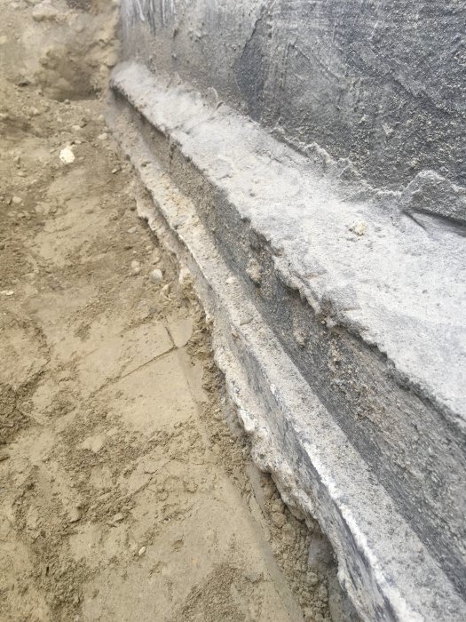 Grävd grund vid hus med ojämn betongsockel och rester av gammalt virke.