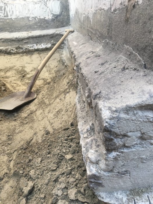 Grävd grund med synliga ojämnheter och utstick i betongfundamentet nära spade och jord.