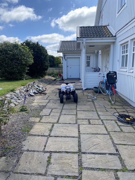 Sjunkna stenplattor på en gångväg med smuts och mossa, framför ett vitt hus med cyklar och verktyg.