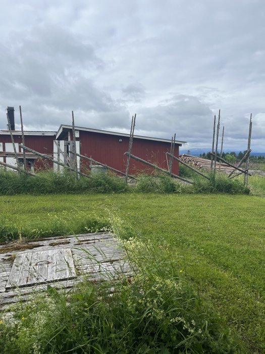 Röd byggnad med vedhög och oavslutad staketkonstruktion på en gräsmatta, potentiell plats för hundgård nära en brunn.