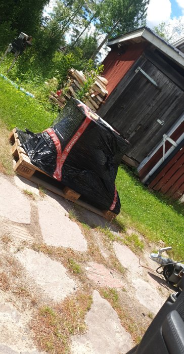 Paket med klinkerplattor inslagna i svart plast på en pall, levererade utomhus.