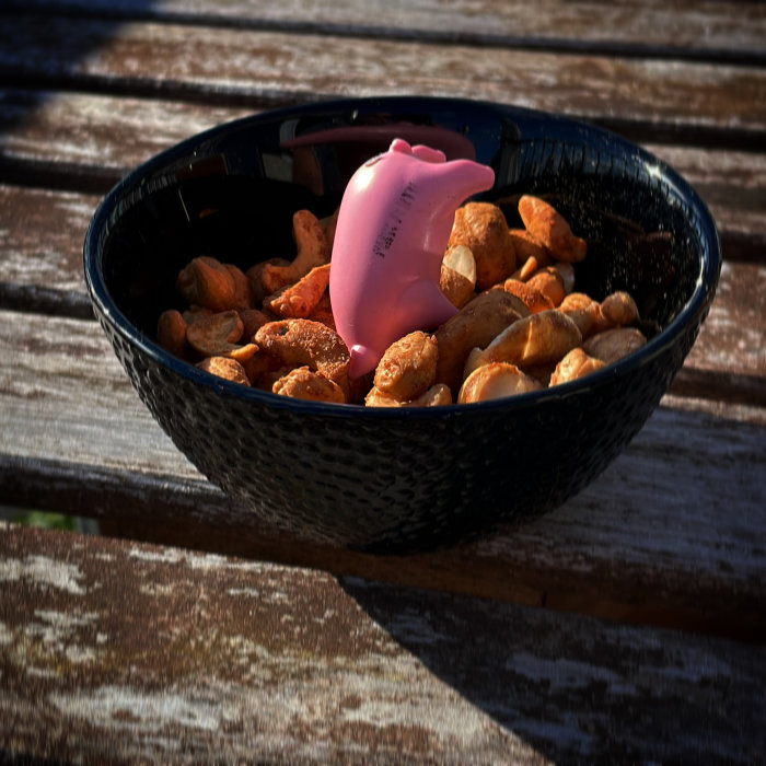 En skål med rökta och jalapeno cashewnötter och en rolig rosa plastgris på en träbänk.