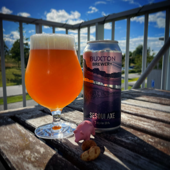 Ölglas med orangefärgad dryck, burk från Buxton Brewery och en liten figur samt cashewnötter på ett träbord utomhus.