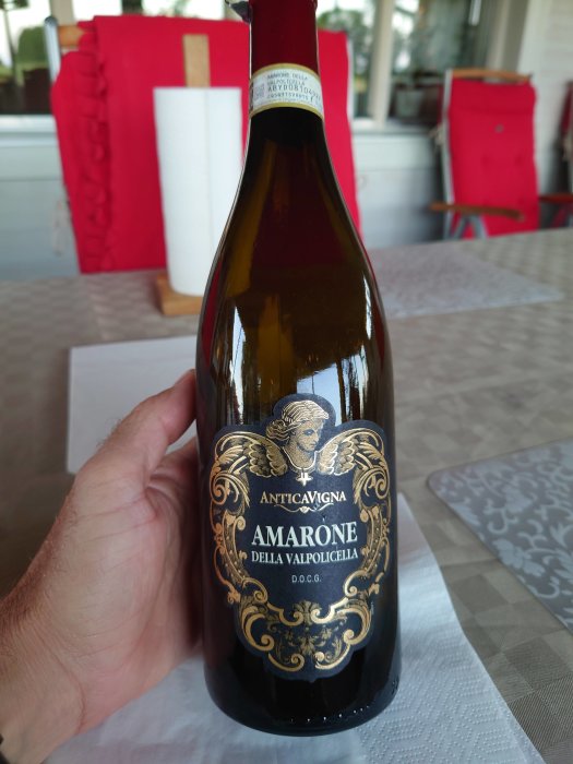 Hand håller en flaska Amarone vin på ett bord, med mat- och vinutrustning i bakgrunden.
