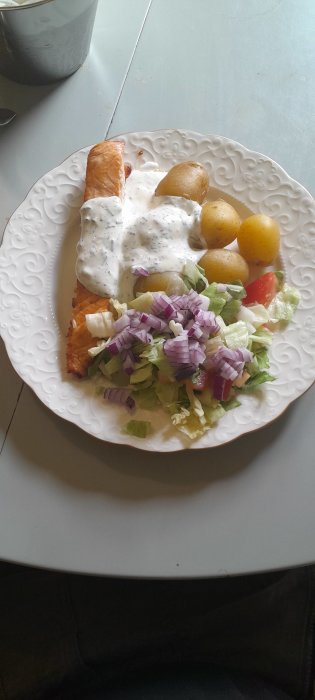 En tallrik med lax, potatis, sallad och sås serverad på ett vitt mönstrat fat.