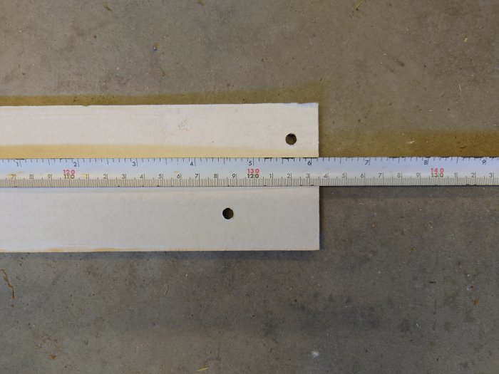 Måttband ligger över två träplattor med förborrade hål markerade för montering, ligger på en betongyta. CC90 avstånd mellan hålen.