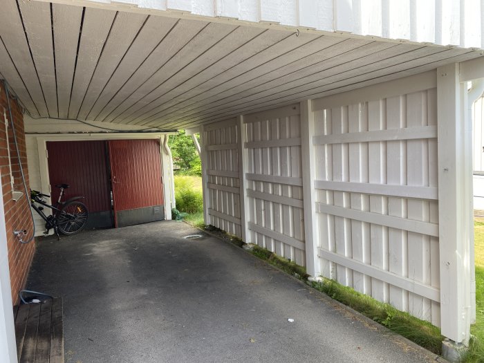 Vit träpanel på väggar och tak i en liten carport med röd garageport och en parkerad cykel.