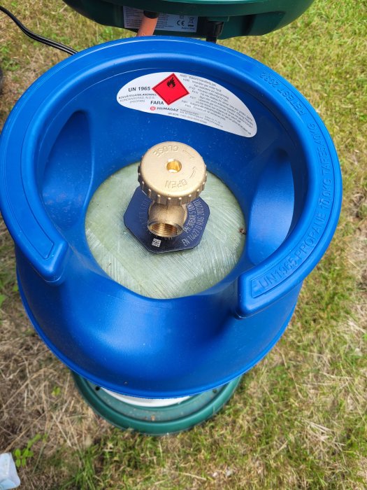 Översikt av en blå gasflaska mot gräset med en mässingsventil och etiketter som visar säkerhetsinformation.
