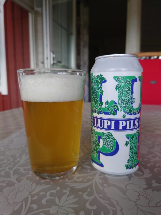 Ölglas med ljust öl och en burk med texten "LUPI PILS" på ett bord framför röd vägg.