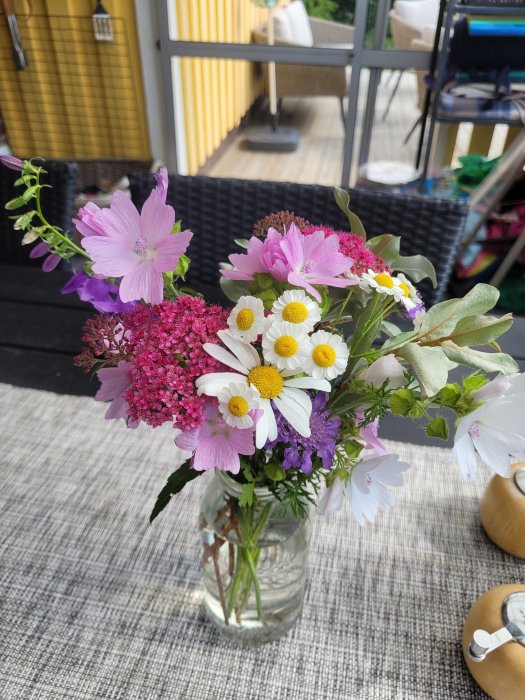 Blommor i olika färger arrangerade i en glasburk på ett utebord med terrass i bakgrunden.