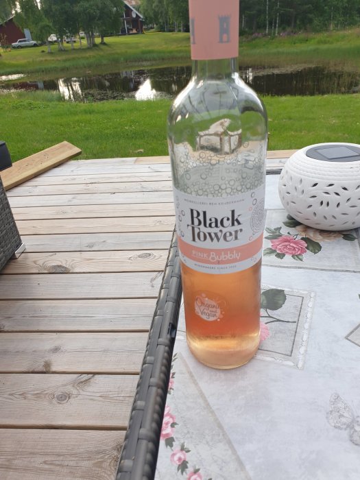 Flaska av Black Tower Pink Bubbly på ett träbord utomhus med damm och grönska i bakgrunden.