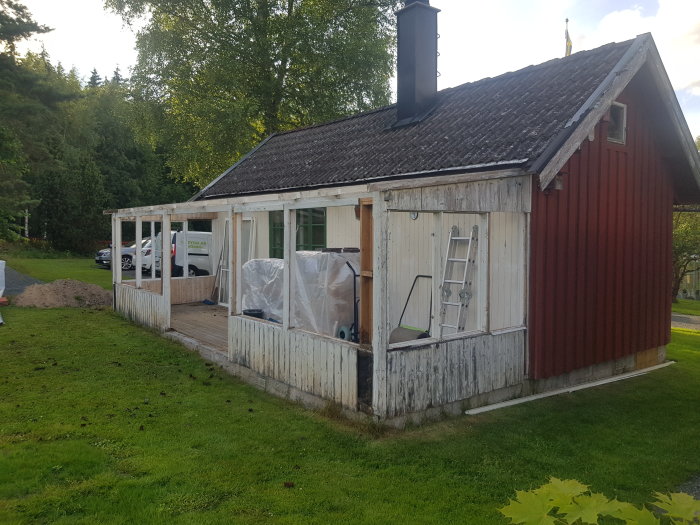 Röd gäststuga med påbörjad renovering av altan, vita plankor delvis avtäckta, stege mot väggen.