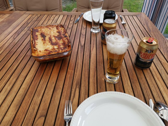 En ugnsform med lasagne på ett träbord, omgiven av ett ölglas, en ölburk och ett tomt tallrik.