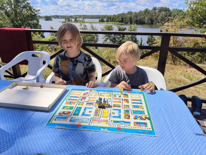 Två barn spelar brädspel utomhus vid ett bord med sjöutsikt.