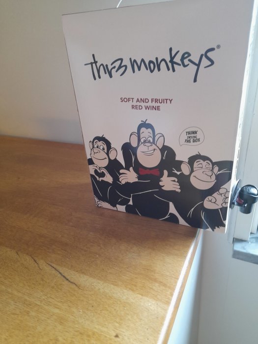 En box med mjuk och fruktig rödvin "Three Monkeys", står på en träyta vid ett fönster.