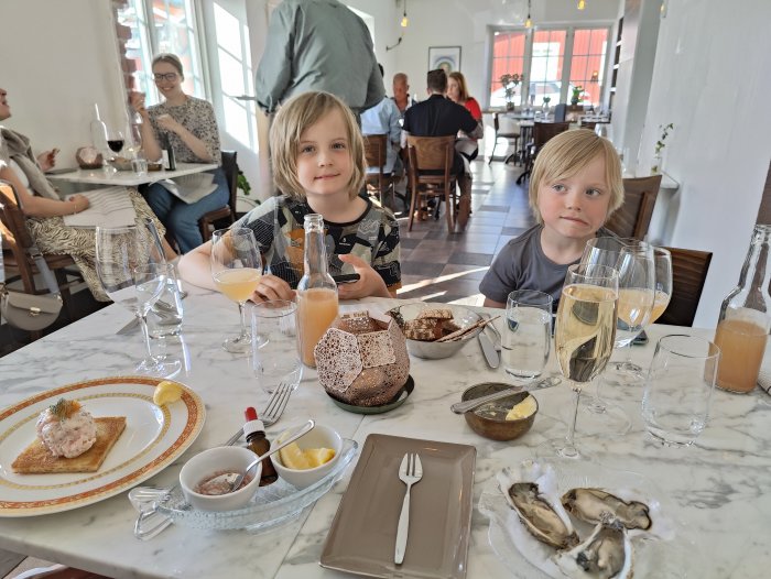 Två barn sitter vid ett dukat restaurangbord med glas och maträtter, mobil ej synlig.