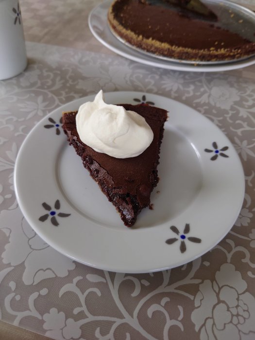 En bit kladdkaka med grädde på ett vitt fat med bakgrund av en hel kladdkaka och en kaffekanna.