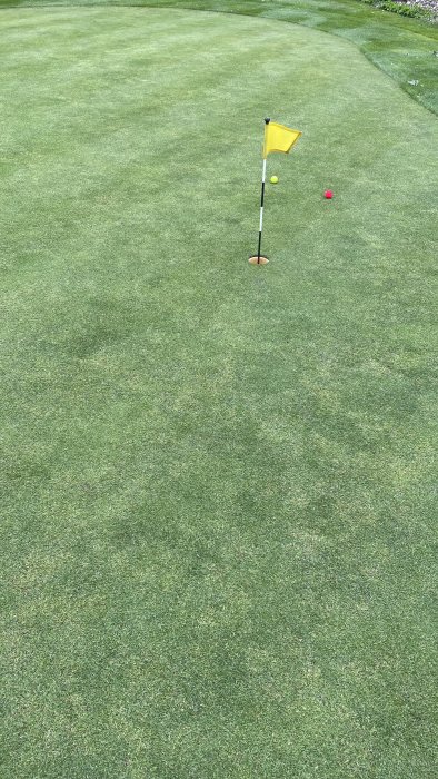 Välskött gräsmatta med golfhål och gul flagga, två golfbollar nära hålet.