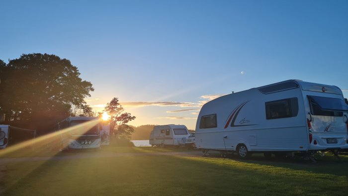 Husvagnar parkerade på en campingplats i solnedgången med träd och vatten i bakgrunden.