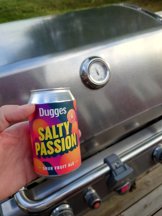 Hand håller en burk Dugges Salty Passion sour fruit ale framför en grill med termometer.