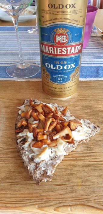 Knäckebröd med smör och stekta kantareller framför en burk Mariestads Old Ox pilsner på ett bord.