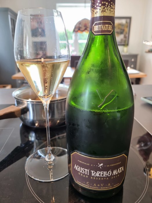 En flaska Brut Nature Cava och ett glas fyllt med mousserande vin, på ett köksbord.