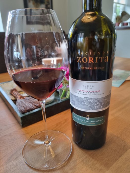 Ett glas rödvin och en flaska Hacienda Zorita Natural Reserve Syrah vid ett bord.